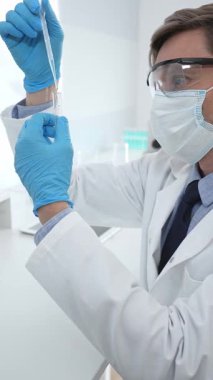 Laboratuvar önlüklü, koruyucu gözlüklü ve mavi eldivenli maskeli erkek bilim adamı modern laboratuardaki test tüplerine mikroskop ve test tüpleri rafı koyuyor. Tıp, sağlık ve bilim konsepti.