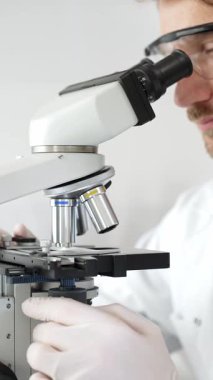 Erkek araştırmacının mikroskopla incelediği örnekler steril laboratuvar ortamında güvenlik gözlüğü takıyor. Tıp, sağlık ve bilim konsepti.