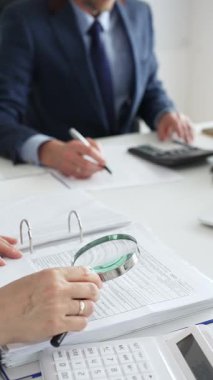 Şirket profesyonelleri iş birliği yapıyor, büyüteç kullanarak finansal evrakları inceliyor, ekip toplantısı sırasında verileri hesaplıyor ve belgeliyor. İş dünyasında denetim ve vergi teması
