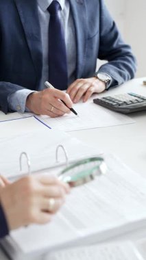 Şirket profesyonelleri iş birliği yapıyor, büyüteç kullanarak finansal evrakları inceliyor, ekip toplantısı sırasında verileri hesaplıyor ve belgeliyor. İş dünyasında denetim ve vergi teması