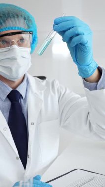 Laboratuvar önlüğü ve eldiveni takan yüz maskesi ve güvenlik gözlüğü takan bilim adamı, laboratuarda bir mikroskop ile mavi sıvıyla test tüpünü inceliyor. Tıp, sağlık ve bilim konsepti