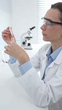 Koruyucu gözlüklü kadın araştırmacı, steril bilimsel çalışma alanı içinde mikroskopların ve laboratuvar ekipmanlarının yanında duran temiz sıvıyı test tüpünde inceliyor. Tıp ve bilim konsepti.