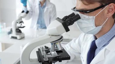 Mikroskop kullanarak maske takan ve eldiven takan bilim adamlarına yakın çekim, modern laboratuvarda araştırmalar yürütmek ve arka planda çalışmak. Tıp, sağlık ve bilim konsepti