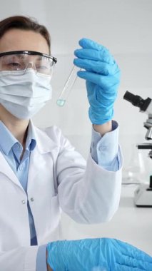 Maske takan, koruyucu gözlük takan ve eldiven takan kadın bilim adamı modern laboratuardaki tıbbi araştırma sırasında mavi sıvıyı dikkatlice inceliyor. Tıp, sağlık ve bilim konsepti