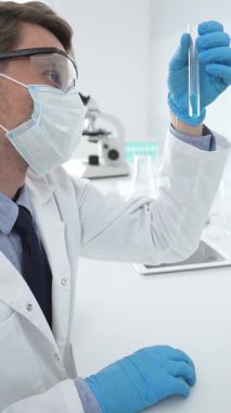 Koruyucu giysi giyen bilim araştırmacısı modern laboratuvar çalışma alanında mavi sıvı numunesini test tüpünden inceliyor. Tıp, sağlık ve bilim konsepti