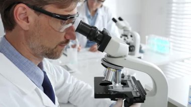 Koruyucu gözlük takan bilim adamı mikroskop altında araştırma ve analiz yapıyor. Arka planda test tüpleri ile modern bir laboratuvarda. Tıp, sağlık ve bilim konsepti