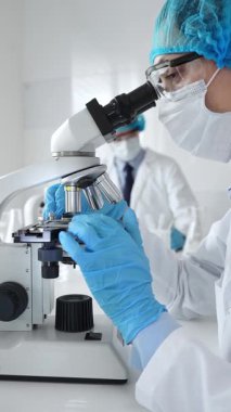 Maske takan ve mavi güvenlik donanımı takan kadın bir bilim adamı araştırma için mikroskop kullanırken erkek bir meslektaşı arka planda çalışıyor. Tıp, sağlık ve bilim konsepti.