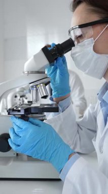 Kadın araştırmacı mikroskop ile örnek analiz ediyor, koruyucu teçhizat takılı, steril araştırma ortamında gelişmiş laboratuvar ekipmanlarının yanında çalışan bir meslektaşı. Tıp, sağlık ve bilim konsepti