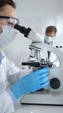 Koruyucu gözlük takan kadın bilim adamı mikroskop kullanarak modern laboratuvarda deney tüpleri ve arka planda erkek bilim adamı ile araştırma yapıyor. Tıp, sağlık ve bilim konsepti