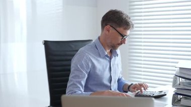 Mavi gömlekli ve gözlüklü profesyonel bir muhasebeci ofis ortamında mali raporları incelerken fatura hesaplıyor ve faturaları yönetiyor. İş dünyasında denetim ve vergi teması.