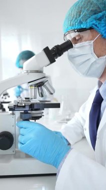 Koruyucu maske, şapka ve eldiven takan kıdemli erkek bilim adamı modern bir laboratuvarda mikroskop altında örnekleri inceliyor. Tıp, sağlık ve bilim konsepti.