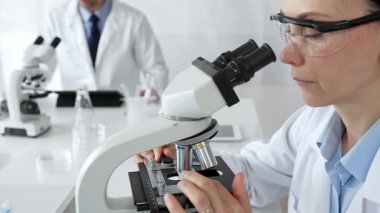 Koruyucu gözlük takan kadın bilim adamı mikroskop kullanarak modern laboratuvarda deney tüpleri ve arka planda erkek bilim adamı ile araştırma yapıyor. Tıp, sağlık ve bilim konsepti