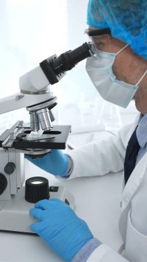 Koruyucu maske takan kıdemli erkek bilim adamı laboratuarda mikroskop kullanarak tıbbi araştırma ve analiz yapıyor. Tıp, sağlık ve bilim konsepti.