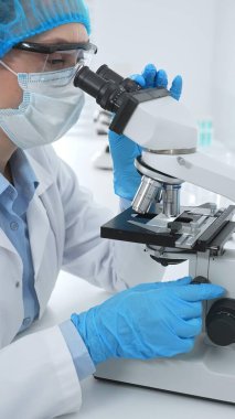 Kadın bilim adamı mikroskop ile numune analiz ediyor, tıbbi araştırma sırasında koruyucu laboratuvar giysileri giyiyor. Tıp, laboratuvar ve bilim konsepti.