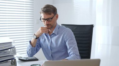 Ciddi bir erkek muhasebeci vergi belgelerini incelerken, dizilmiş dosyaların yanında hesap makinesi kullanarak, açık mavi gömlekli, siyah ofis koltuğunda oturarak düşünüyor. Ticarette denetim ve vergiler.