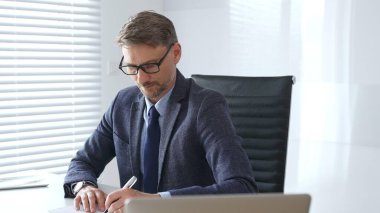 Kıdemli işadamı takım elbise, kuyruk ve siyah gözlük takarak masasındaki parlak, modern bir ofiste profesyonelliği gösteren bir belgeye konsantre oluyor. İş adamları konsepti.