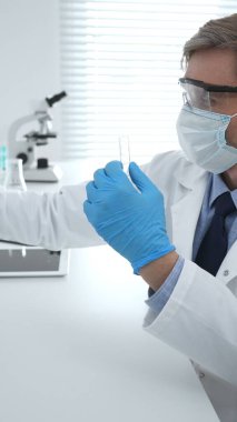 Laboratuvar önlüklü, koruyucu gözlüklü ve mavi eldivenli maskeli erkek bilim adamı modern laboratuardaki test tüplerine mikroskop ve test tüpleri rafı koyuyor. Tıp, sağlık ve bilim konsepti.