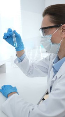 Laboratuvar önlüğü giyen ve koruyucu gözlük takan kadın bilim adamı modern laboratuarda mikroskop ve test tüpleri askısı ile test tüpünü temiz sıvıyla analiz ediyor. Tıp, sağlık ve bilim konsepti