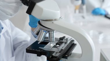 Mavi eldiven ve maske takan bilim adamları, laboratuvar ortamında modern bir mikroskop kullanarak araştırma ve analiz yapıyorlar. Tıp, sağlık ve bilim konsepti