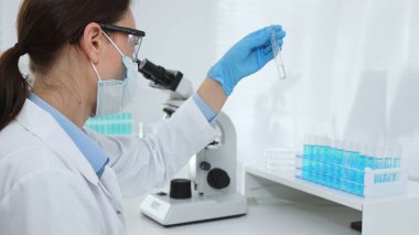 Koruyucu maske takan dişi araştırmacı ve mavi eldiven test tüpünde sıvıyı inceliyor, laboratuvarda mikroskop ve laboratuvar ekipmanlarının yanında oturuyor. Tıp, sağlık ve bilim konsepti.