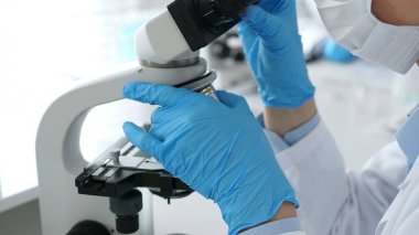 Mavi eldiven ve maske takan bilim adamları, laboratuvar ortamında modern bir mikroskop kullanarak araştırma ve analiz yapıyorlar. Tıp, sağlık ve bilim konsepti