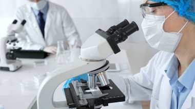 Maske takan ve mavi güvenlik donanımı takan kadın bir bilim adamı araştırma için mikroskop kullanırken erkek bir meslektaşı arka planda çalışıyor. Tıp, sağlık ve bilim konsepti.