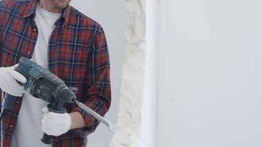 Kırmızı kareli gömlek ve koruyucu beyaz eldiven giyen erkek inşaat işçisi, yenileme projesi sırasında beyaz duvarı döner çekiçle deliyor ve toz üretiyor, yakın görüş.