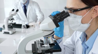 Mavi maskeli ve eldivenli bir kadın bilim adamı arka planda çalışan bir meslektaşı steril bir laboratuvarda mikroskop kullanıyor ve bilimsel araştırmalar yürütüyor. İlaç, sağlık hizmetleri ve
