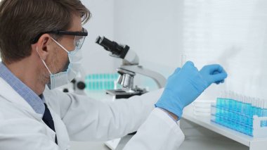 Laboratuvar önlüklü, koruyucu gözlüklü ve mavi eldivenli maskeli erkek bilim adamı modern laboratuardaki test tüplerine mikroskop ve test tüpleri rafı koyuyor. Tıp, sağlık ve bilim konsepti.