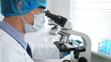 Koruyucu maske, şapka ve eldiven takan kıdemli erkek bilim adamı modern bir laboratuvarda mikroskop altında örnekleri inceliyor. Tıp, sağlık ve bilim konsepti.