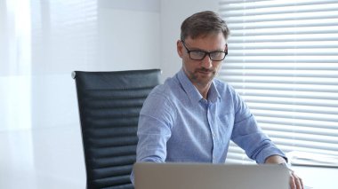 Mavi parlak gömlek ve siyah gözlük takan işadamı, parlak ve modern bir ofiste çalışırken, profesyonellik ve verimliliğini gösteriyor. İş adamları konsepti.