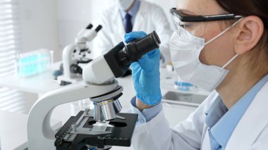 Mavi maskeli ve eldivenli bir kadın bilim adamı arka planda çalışan bir meslektaşı steril bir laboratuvarda mikroskop kullanıyor ve bilimsel araştırmalar yürütüyor. İlaç, sağlık hizmetleri ve