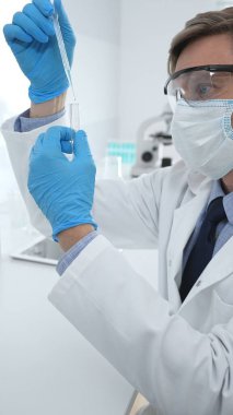 Laboratuvar önlüklü, koruyucu gözlüklü ve mavi eldivenli maskeli erkek bilim adamı modern laboratuardaki test tüplerine mikroskop ve test tüpleri rafı koyuyor. Tıp, sağlık ve bilim konsepti.