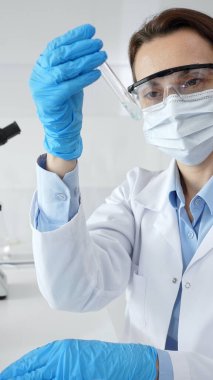 Steril laboratuvar ortamında tıbbi deney sırasında koruyucu giysi giyerken mavi sıvıyı analiz eden deney tüpü tutan kadın araştırmacı. Tıp ve bilim konsepti.