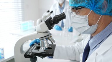 Koruyucu maske, şapka ve eldiven takan kıdemli erkek bilim adamı modern bir laboratuvarda mikroskop altında örnekleri inceliyor. Tıp, sağlık ve bilim konsepti.