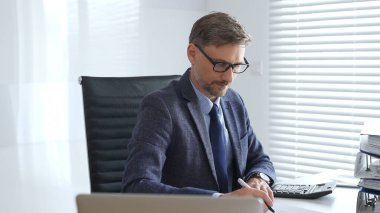 Takım elbiseli, kravatlı ve gözlüklü olgun girişimci masada oturuyor, notlar yazıyor ve modern, parlak ofiste hesap makinesi kullanıyor. Ticarette denetim ve vergiler.