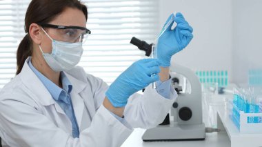 Yüz maskesi takan ve mavi eldiven takan kadın bir bilim adamı modern bir laboratuarda bir deney tüpüne dikkatlice sıvı bırakıyor. Tıp ve bilim konsepti.