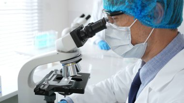 Koruyucu maskeli, şapkalı ve eldivenli erkek bilim adamı steril laboratuvarda mikroskop altında örnekler üzerinde çalışıyor. Tıp, sağlık ve bilim kavramı.