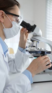 Maskeli profesyonel kadın bilim adamı mikroskop ile numune inceleyerek steril laboratuvar ortamında meslektaşlarıyla birlikte araştırma yapıyor. Tıp, sağlık ve bilim konsepti.