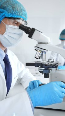 Koruyucu maske, şapka ve eldiven takan kıdemli erkek bilim adamı modern bir laboratuvarda mikroskop altında örnekleri inceliyor. Tıp, sağlık ve bilim konsepti.