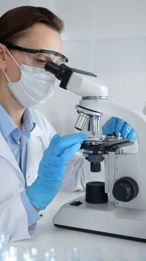 Maske takan ve mavi eldiven giyen kadın bilim adamı mikroskop kullanarak arka planda çalışan bir meslektaşla modern laboratuvarda bilimsel araştırmalar yapıyor. Tıp, sağlık ve bilim konsepti.
