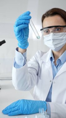 Steril laboratuvar ortamında tıbbi deney sırasında koruyucu giysi giyerken mavi sıvıyı analiz eden deney tüpü tutan kadın araştırmacı. Tıp ve bilim konsepti.
