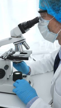 Koruyucu maske takan kıdemli erkek bilim adamı laboratuarda mikroskop kullanarak tıbbi araştırma ve analiz yapıyor. Tıp, sağlık ve bilim konsepti.