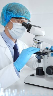 Koruyucu maske, şapka ve eldiven takan kıdemli erkek bilim adamı modern bir laboratuvarda mikroskop altında örnekleri inceliyor. Tıp, sağlık ve bilim konsepti.