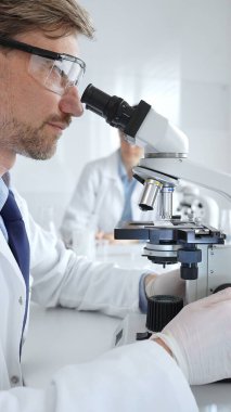 Koruyucu gözlük takan bilim adamı mikroskop altında araştırma ve analiz yapıyor. Arka planda test tüpleri ile modern bir laboratuvarda. Tıp, sağlık ve bilim konsepti