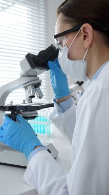 Maske takan ve mavi eldiven giyen kadın bilim adamı mikroskop kullanarak arka planda çalışan bir meslektaşla modern laboratuvarda bilimsel araştırmalar yapıyor. Tıp, sağlık ve bilim konsepti.