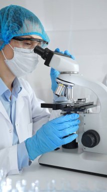 Maske takan ve mavi güvenlik donanımı takan kadın bir bilim adamı araştırma için mikroskop kullanırken erkek bir meslektaşı arka planda çalışıyor. Tıp, sağlık ve bilim konsepti.