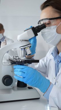 Maske takan ve mavi eldiven giyen kadın bilim adamı mikroskop kullanarak arka planda çalışan bir meslektaşla modern laboratuvarda bilimsel araştırmalar yapıyor. Tıp, sağlık ve bilim konsepti.