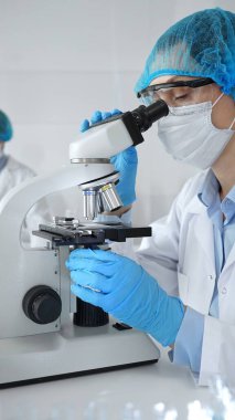 Bilinmeyen bilim adamı mavi güvenlik donanımı takıyor. Araştırma için mikroskop kullanıyor. Erkek bir meslektaşı arka planda çalışıyor. Tıp, sağlık ve bilim konsepti.