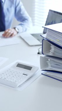 Ofis çalışanları mali raporları analiz ediyor ve ön planda evrak dolu mavi dosyalarla dolu beyaz bir masada otururken belgeleri imzalıyorlar. İş dünyasında denetim ve vergi teması